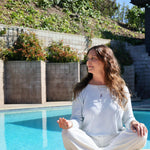 Maya Brenner wearing a yellow gold Bold Link Chain, the Healing Retreat Necklace, multiple rings and bracelets, seated outdoors with a pool in the background.