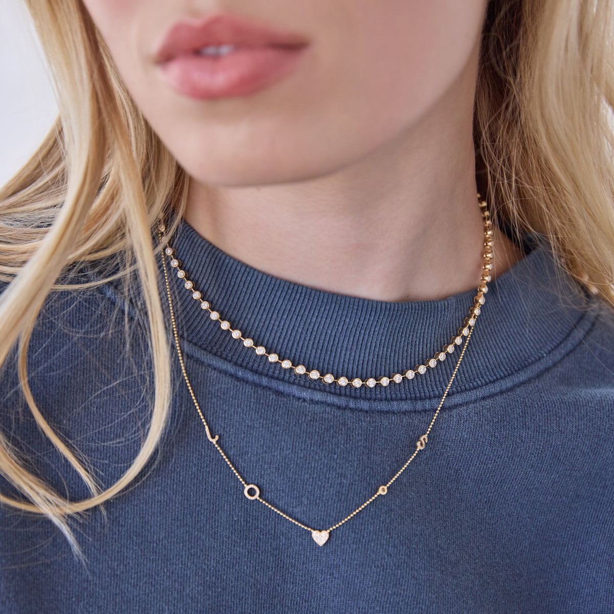 Close-up of a woman's neck wearing The Ultimate Diamond Necklace and a Custom Gold Necklace - 5 Letters, with a blue jumper and blonde hair. 