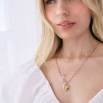 A blonde woman wearing an Element Short Link Chain, a Custom Gold Necklace - 5 Letters, The Love Retreat Necklace and The Peony Bouquet Necklace with a Diamond Heart Locket Charm, styled in a white top against a white background.