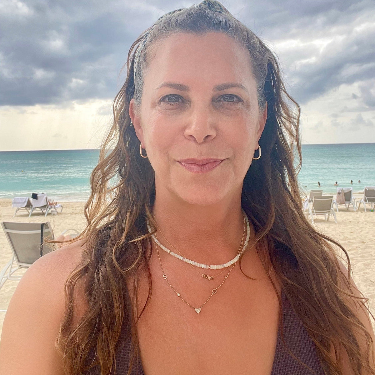 Maya Brenner wearing an Endless Summer Necklace - Pink Sands, a Petite Nameplate Necklace with 'maya', a Custom Gold Necklace - 5 Letters and Oval Hoops, with the beach as the background.