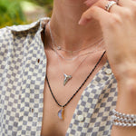 Close-up of a neck wearing a Custom Gold Necklace - 2 Letters, an Element Long Link Chain, and a Sharktooth Necklace, all in white gold, along with a Black Rhodium Antique Bead Chain with a Moonstone Crescent Charm, as well as Rolo Bracelets and Bouquet Rings, styled in a checkered shirt with a green blurred background.