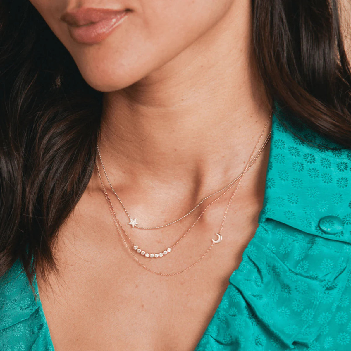A close-up of a woman's neck wearing two 14k Gold Asymmetrical Charm Necklaces with star and moon charms, along with a Diamond Arc Layering Necklace in rose or white gold, styled in a turquoise blue shirt.