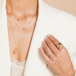 Close-up of a woman's décolletage wearing several Custom Gold Necklaces with charms and a Zodiac Necklace - Scorpio, with her hand wearing a Paris Ring, a Link Ring, and two Pavé Diamond Rings, with a cream lace top and a white blazer against a neutral background.