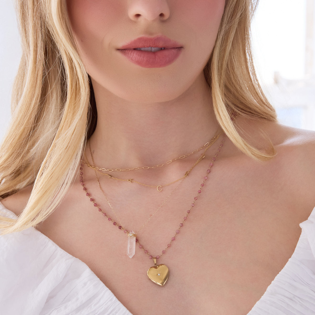 A close-up of a blonde woman's décolletage wearing an Element Short Link Chain, a Custom Gold Necklace - 5 Letters, The Clarity Retreat Necklace and The Peony Bouquet Necklace with a Diamond Heart Locket Charm, with a white top and white background. 
