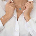 Close-up of a woman's decellotage wearing several The Lily Bouquet Necklaces with an Etched Turquoise Heart Charm, with her hands wearing The Lily Bouquet Bracelet, a Baroque White Pearl Bangle and a Perla Pinky Ring, in a white shirt. 