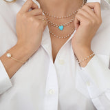 Close-up of a woman's decellotage wearing several The Lily Bouquet Necklaces with an Etched Turquoise Heart Charm, with her hands wearing The Lily Bouquet Bracelet, a Baroque White Pearl Bangle and a Perla Pinky Ring, in a white shirt. 