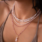 Close-up of a woman wearing multiple Endless Summer Necklaces in Pink Sands and The Peony Bouquet Necklace with a Balance Crystal Charm in Rose Quartz, styled in a beige strap top. 
