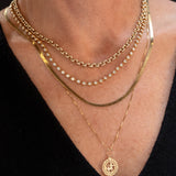 Close-up of a neck wearing a Rolo Necklace, the Ultimate Diamond Necklace, a Sequence Chain and a Zodiac Necklace - Sagittarius, all in yellow gold, with a black top. 