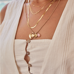 Close-up of a woman wearing a Custom Gold Necklace – 2 Letters, Overboard Layering Chain, Element Short Link Chain, Sequence Chain and Round Curb Chain with a Pink Opal Tear Drop Charm, Heart Locket Charm, Diamond Heart Locket Charm and Engraved Heart Charm, styled in a white linen top against a blurred outdoor background.