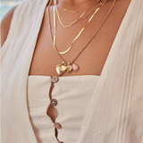 Close-up of a woman wearing a Custom Gold Necklace – 2 Letters, Overboard Layering Chain, Element Short Link Chain, Sequence Chain and Round Curb Chain with a Pink Opal Tear Drop Charm, Heart Locket Charm, Diamond Heart Locket Charm and Engraved Heart Charm, styled in a white linen top against a blurred outdoor background.