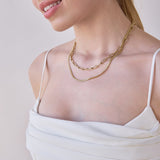 Close-up of a woman's décolletage wearing a Bold Link Chain and a Chic Cuban Curb Chain, both in yellow gold, with a white strap top against a light grey background.