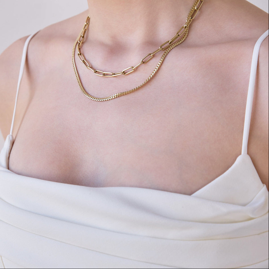Close-up of a woman's décolletage wearing a Bold Link Chain and a Chic Cuban Curb Chain, both in yellow gold, with a white strap top against a light grey background.