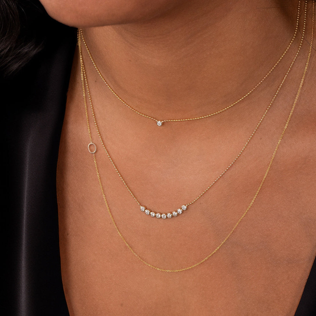 Close-up of a neck wearing a Single Diamond Layering Necklace, a Diamond Arc Layering Necklace and a 14k Gold Asymmetrical Letter Necklace - O all in yellow gold, with a black silk top.