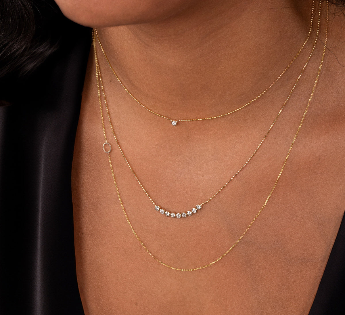 Close-up of a neck wearing a Single Diamond Layering Necklace, a Diamond Arc Layering Necklace and a 14k Gold Asymmetrical Letter Necklace - O all in yellow gold, with a black silk top. 