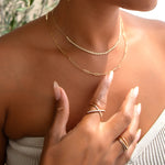 Close-up of a woman’s décolletage wearing the Everyday Diamond Necklace layered with an Element Long Link Chain, with her hand wearing a Criss-Cross Diamond Daze Ring and a Criss-Cross Diamond Duo Ring, styled with a strapless neutral top against a blurred leafy background.