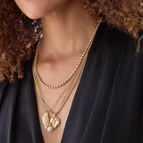 Close-up of a person's décolletage wearing the Ultimate Diamond Necklace, a Box Chain with a Diamond Heart Locket Charm, an Element Long Link Chain with the 20th Anniversary Leaf Pendant, and a Perla Necklace, all in yellow gold, with a black top and brown curly hair.
