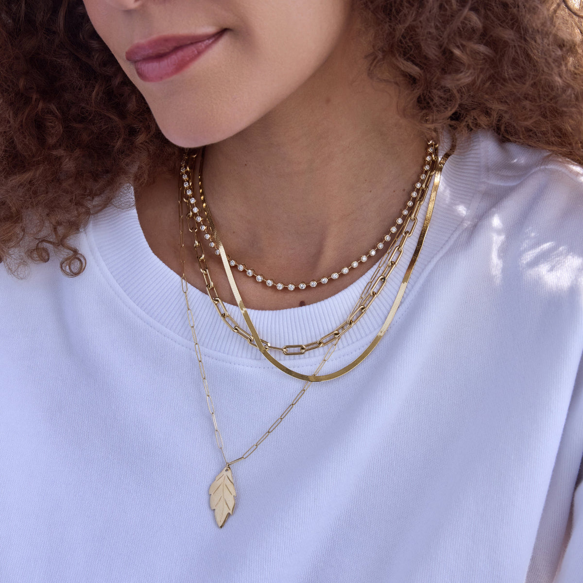 Close-up of a woman's neck wearing the Ultimate Diamond Necklace, a Bold Link Chain, a Sequence Chain, and an Element Long Link Chain with a 20th Anniversary Leaf Pendant, styled in a white T-shirt with curly hair.