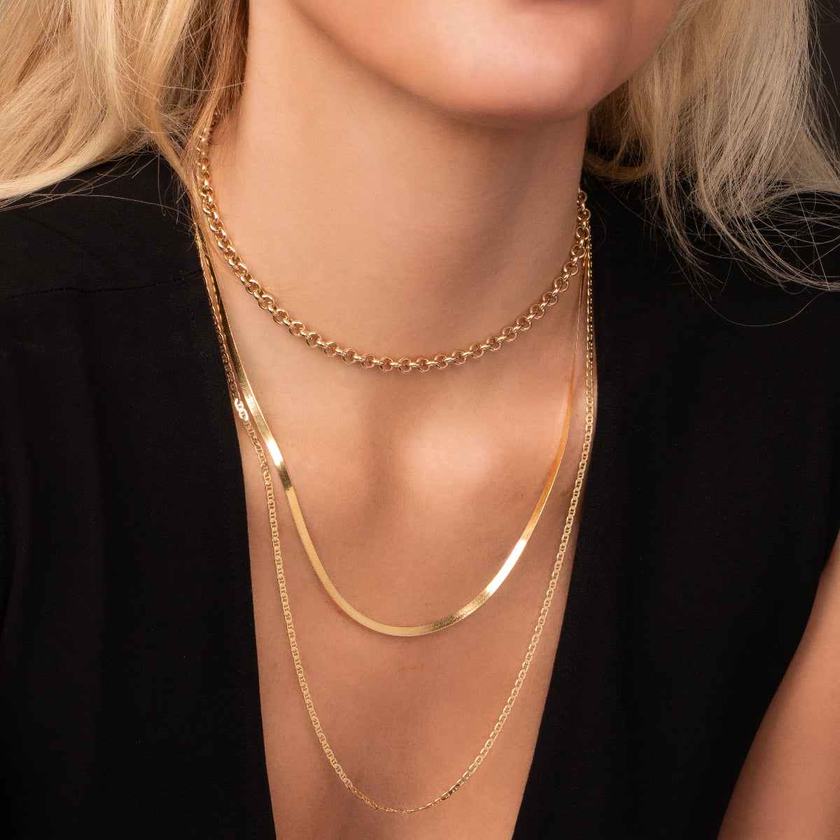 Close-up of a blonde woman’s décolletage wearing a yellow gold Rolo Necklace, a Sequence Chain and an Overboard Layering Necklace, with a black top.