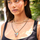 A woman wearing several necklaces mainly in yellow gold, including an Element Long Link Chain, a Rolo Necklace, a Custom Gold Necklace - 4 Letters, a Zodiac Necklace - Gemini, a Black Rhodium Antique Bead Chain and a Box Chain with several charms, including an Etched Turquoise Heart Charm, a Diamond Heart Locket Charm, a Balance Crystal Charm and a Small Abalone Cabochon Charm, along with Solid Eclipse Earrings - Small, styled in a black top against a green leafy tropical background.