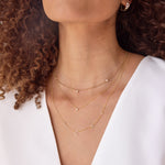 Close-up of a woman’s neck wearing a yellow gold Triple Diamond Layering Necklace, a Single Diamond Layering Necklace, and a Custom 5-Letter Gold Necklace, along with a Tres Pearl Earring and a Pavé Huggie, paired with a white top and her brown curly hair visible. 