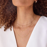 Close-up of a woman’s neck wearing a yellow gold Triple Diamond Layering Necklace, a Single Diamond Layering Necklace, and a Custom 5-Letter Gold Necklace, along with a Tres Pearl Earring and a Pavé Huggie, paired with a white top and her brown curly hair visible. 