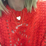 Close-up of blonde woman's décolletage wearing an AMOUR Necklace and a necklace with a heart pendant, both in yellow gold, with a red knit top. 
