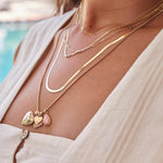 A woman's decellotage wearing a Custom Gold Necklace – 2 Letters, Overboard Layering Chain, Element Short Link Chain, Sequence Chain and Round Curb Chain with a Pink Opal Tear Drop Charm, Heart Locket Charm, Diamond Heart Locket Charm and Engraved Heart Charm, styled in a white linen top against a blurred outdoor background.