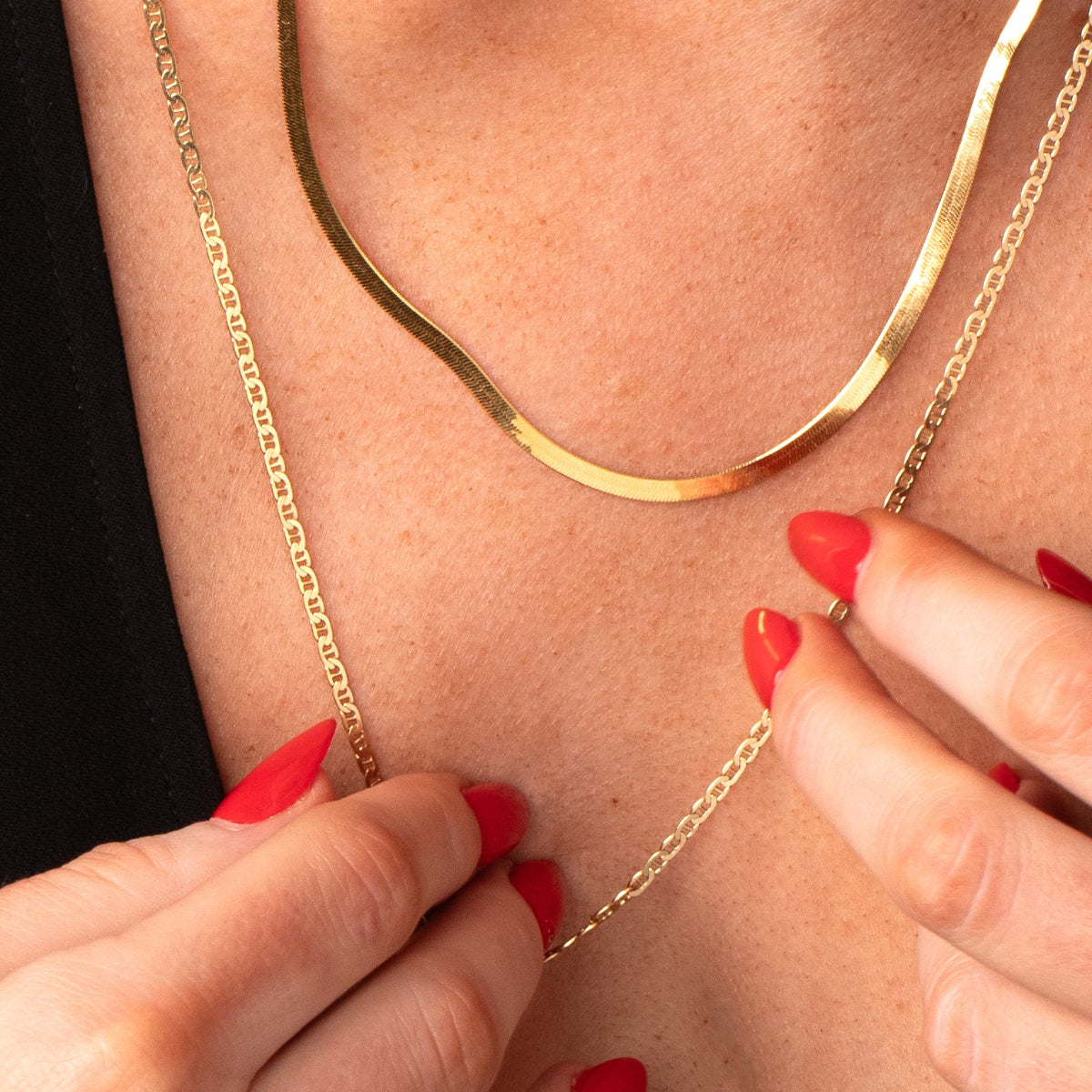 A décolletage wearing a yellow gold Sequence Chain and an Overboard Layering Necklace, with hands featuring red nails. 