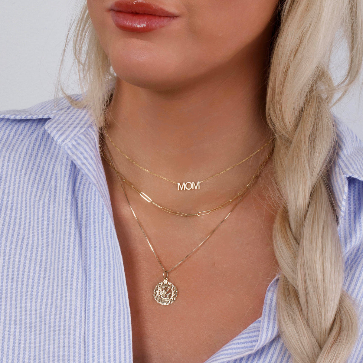 Close-up of a blonde woman's décolletage wearing a MOM Necklace, an Element Long Link Chain and a Zodiac Necklace - Scorpio, all in yellow gold, with a blue and white striped shirt against a light grey background.