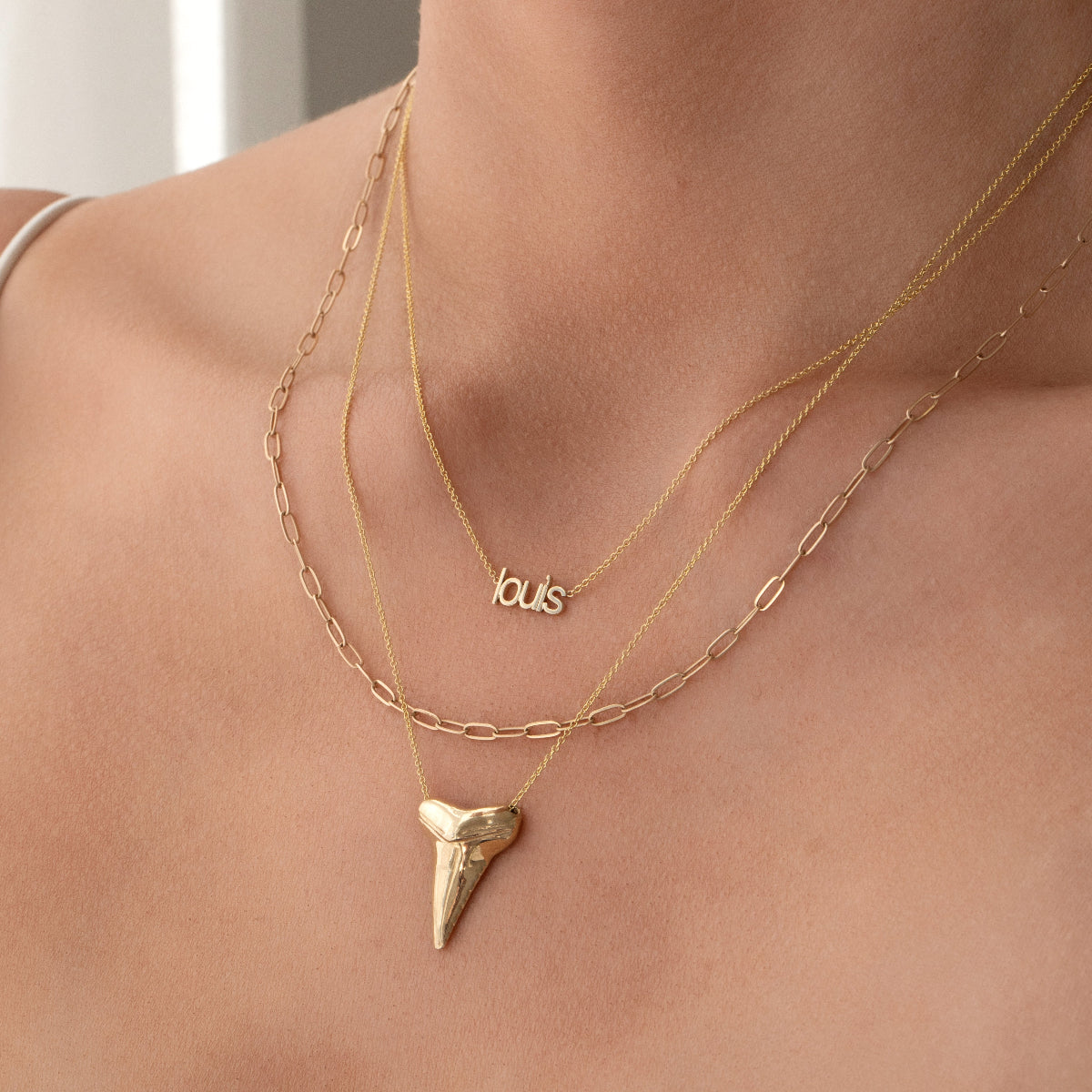 A close-up of a neck wearing a Petite Nameplate Necklace with 'louis', an Element Short Link Chain and a Sharktooth Necklace, all in yellow gold. 