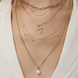 A close-up of a neck wearing a Bold Link Chain, a Diamond Arc Layering Necklace, an Element Long Link Chain with a Diamond Pendant and Zodiac Pendant (Aries) and two Custom Gold Necklaces - 3 Letters, and a Box Chain with a Perla Pendant, all in yellow gold, styled with a cream top. 