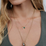 Close-up of a woman wearing a Classic Bead Chain with a Color Pop Pendant in Green (Lab-Grown Emerald) and a Petite Nameplate Pendant with a Pavé Moon Charm, layered with a Box Chain featuring a Color Pop Pendant in Light Blue (Aquamarine) and a Cut-Out Heart Charm, wearing a grey top with her blonde hair visible.