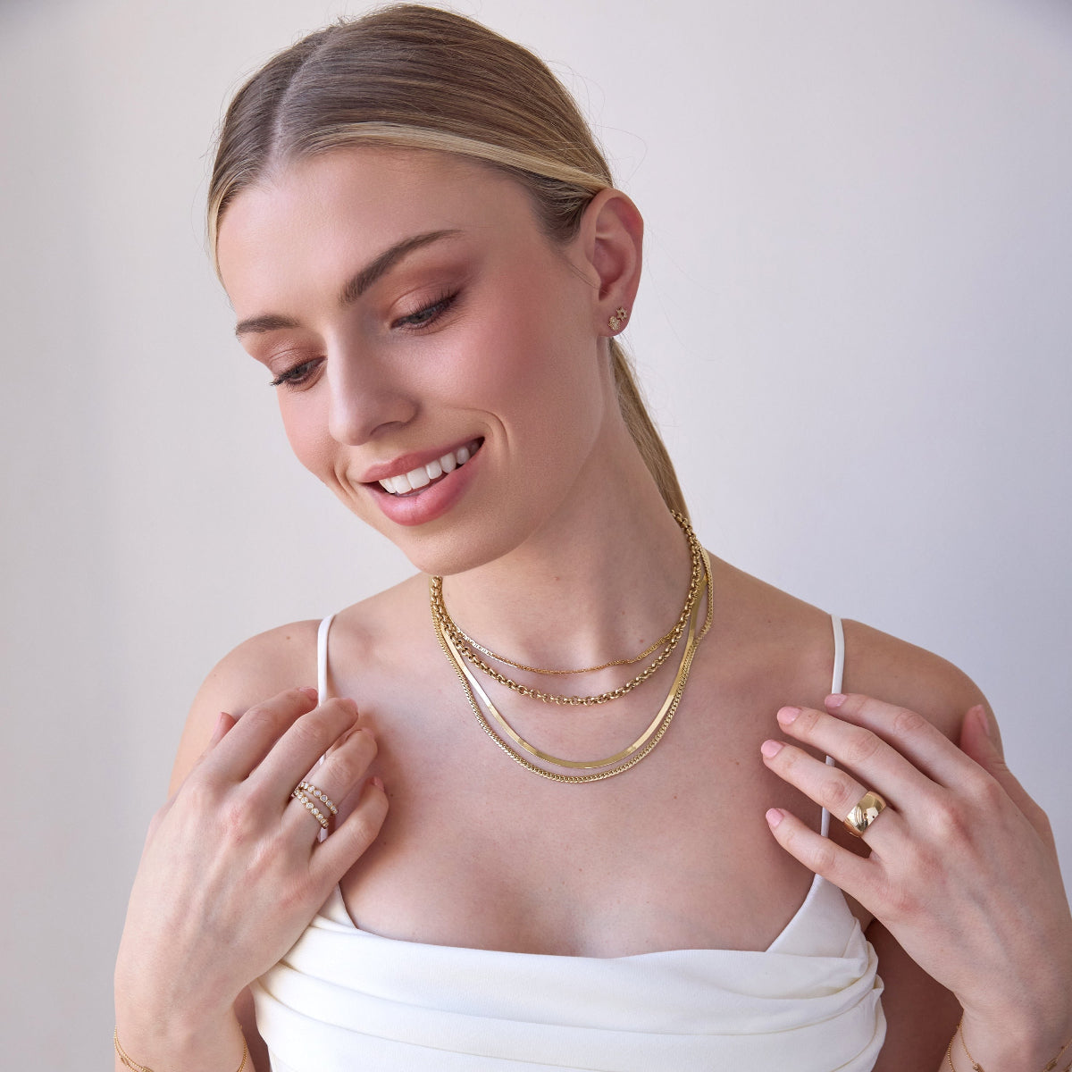 A blonde woman wearing layered yellow gold necklaces, including an Overboard Layering Chain, a Rolo Necklace, a Sequence Chain and a Chic Cuban Curb Chain, along with a Thick Band, two Evermore Rings, a Pavé Charm Stud - Hamsa and a Pavé Charm Stud - Star of David, with a white top against a plain background.