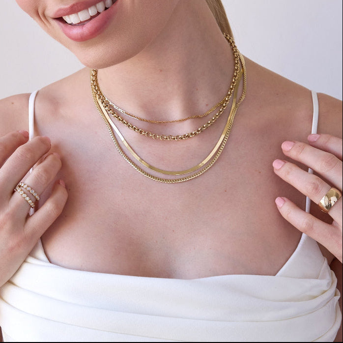 A blonde woman wearing layered yellow gold necklaces, including an Overboard Layering Chain, a Rolo Necklace, a Sequence Chain and a Chic Cuban Curb Chain, along with a Thick Band, two Evermore Rings, a Pavé Charm Stud - Hamsa and a Pavé Charm Stud - Star of David, with a white top against a plain background.
