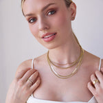 A blonde woman wearing layered yellow gold necklaces, including an Overboard Layering Chain, a Rolo Necklace, a Sequence Chain and a Chic Cuban Curb Chain, along with a Thick Band, two Evermore Rings, a Pavé Charm Stud - Hamsa and a Pavé Charm Stud - Star of David, with a white top against a light grey background.