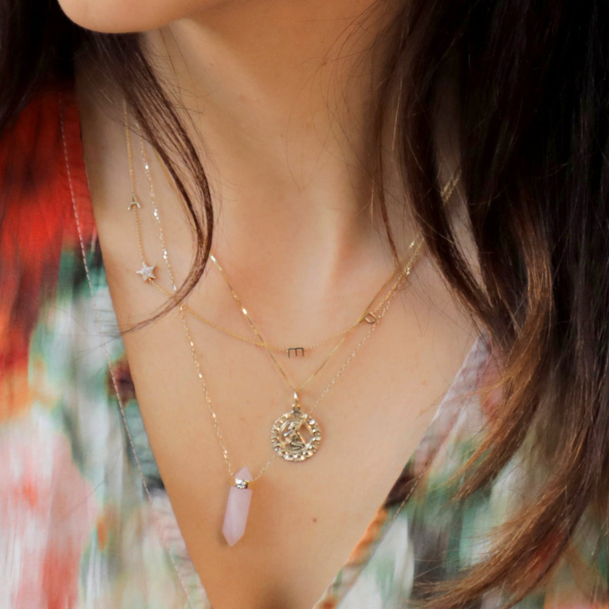 Close-up of a brunette woman's décolletage wearing a Zodiac Necklace - Gemini, The Love Retreat Necklace and a Custom Gold Necklace - 4 Letters with a colourful top.