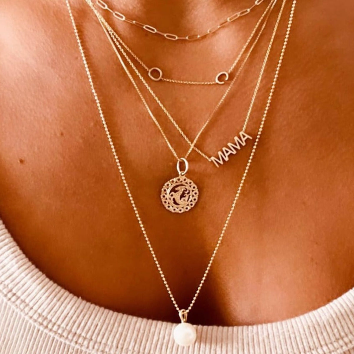 Close-up of a décolletage wearing several gold necklaces - an Element Short Link Chain, a Custom Gold Necklace - 2 Letters, a MAMA Necklace, a Zodiac Necklace - Capricorn and a Perla Necklace. 