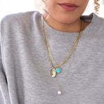 A close-up of a woman wearing a Chic Cuban Curb Chain with a Large Abalone Cabochon Charm, an Element Long Link Chain with a Turquoise Heart Charm and a Perla Pendant on a cable chain necklace, all in yellow gold, with a light grey jumper against a white background.