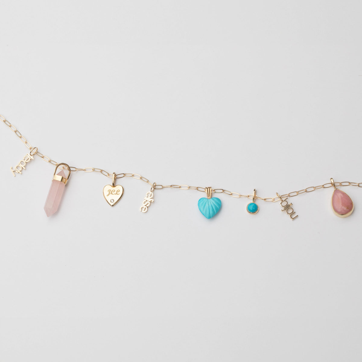 A yellow gold Element Short Link Chain with several pendants and charms, including Petite Nameplate Pendants in 'teddy', 'elise' and 'bijou', a rose quartz Balance Crystal Charm, an Engraved Heart Charm, an Etched Turquoise Heart Charm, a Turquoise Dangle Charm and a Pink Opal Tear Drop Charm, against a light grey background. 