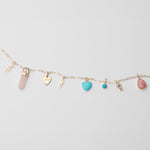 A yellow gold Element Short Link Chain with several pendants and charms, including Petite Nameplate Pendants in 'teddy', 'elise' and 'bijou', a rose quartz Balance Crystal Charm, an Engraved Heart Charm, an Etched Turquoise Heart Charm, a Turquoise Dangle Charm and a Pink Opal Tear Drop Charm, against a light grey background. 