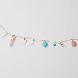 A yellow gold Element Short Link Chain with several pendants and charms, including Petite Nameplate Pendants in 'teddy', 'elise' and 'bijou', a rose quartz Balance Crystal Charm, an Engraved Heart Charm, an Etched Turquoise Heart Charm, a Turquoise Dangle Charm and a Pink Opal Tear Drop Charm, against a light grey background. 