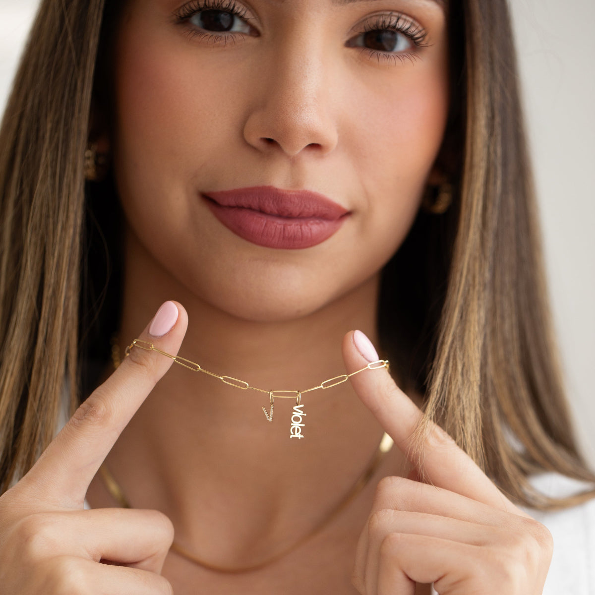 A woman holding up a yellow gold Element Short Link Chain with a Petite Nameplate Pendant ‘violet’ and a Pavé Letter Charm ‘V’, styled with a yellow gold Sequence Chain, earrings, and a white top.