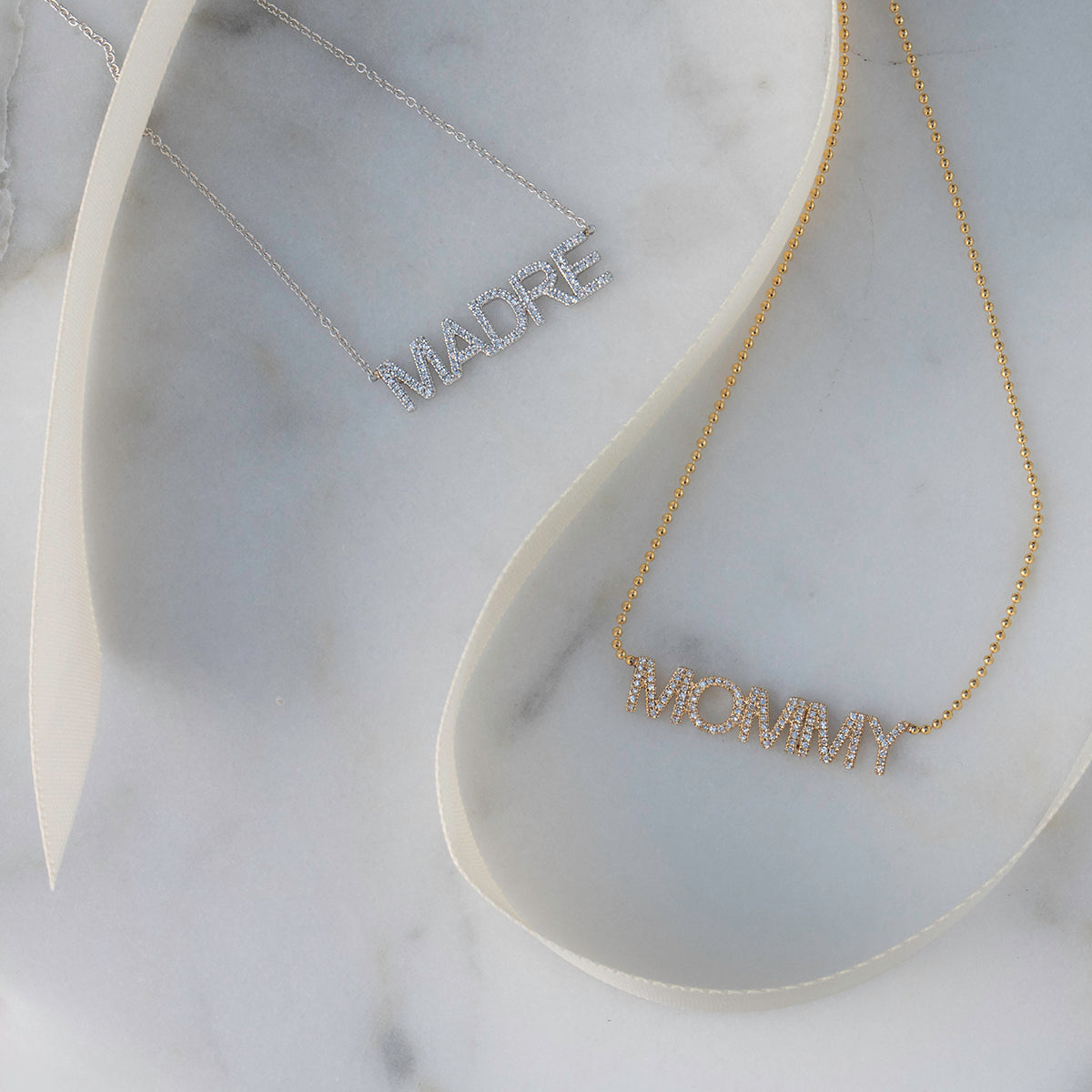A Pavé Madre Necklace in white gold and a Pavé MOMMY Necklace in yellow gold on a white marble background. 