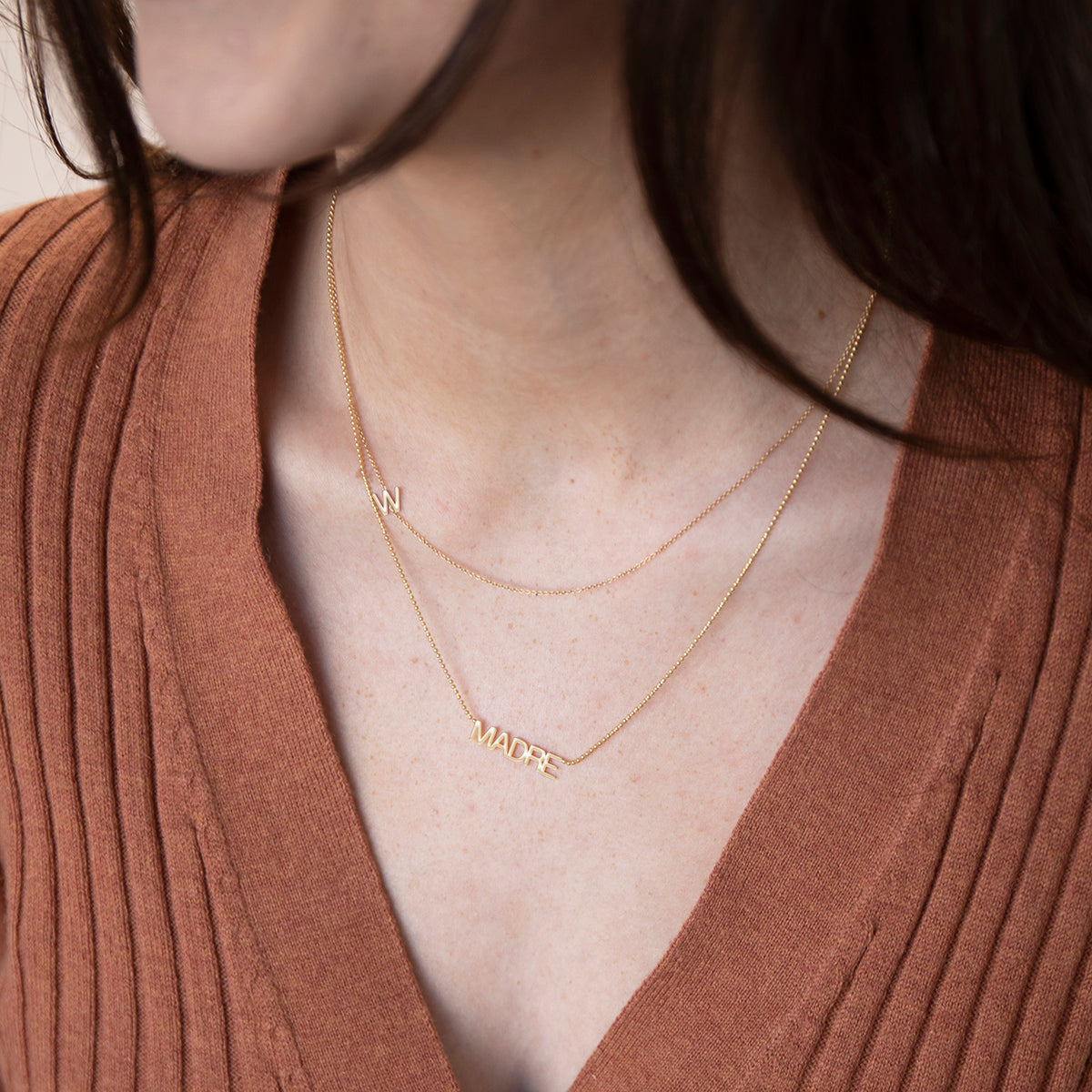 Close-up of a woman's neck wearing a Custom Gold Necklace - 1 Letter (W) and a MADRE Necklace in yellow gold, styled in a burnt orange top with brown hair visible. 