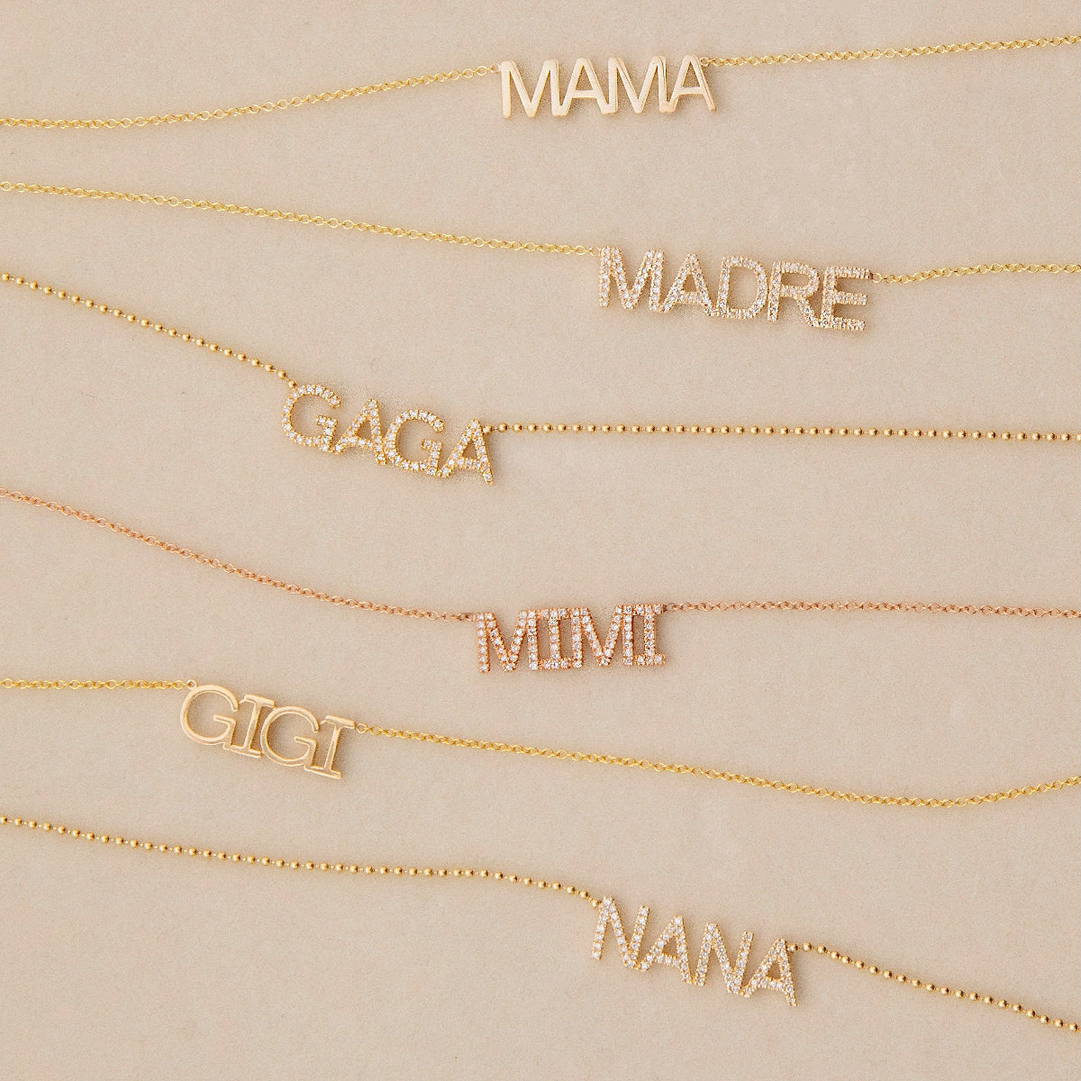 MAMA, Pavé MADRE, Pavé GAGA, Pavé MIMI, GIGI and Pavé NANA Necklaces in yellow or rose gold laid against a light beige background. 
