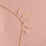 A delicate gold chain necklace laid flat on a dusty pink background, featuring five small dangling nameplates in cursive script reading 'miller', 'willow', 'amefa', 'emma', and 'ava'.