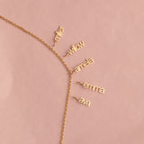 A delicate gold chain necklace laid flat on a dusty pink background, featuring five small dangling nameplates in cursive script reading 'miller', 'willow', 'amefa', 'emma', and 'ava'.