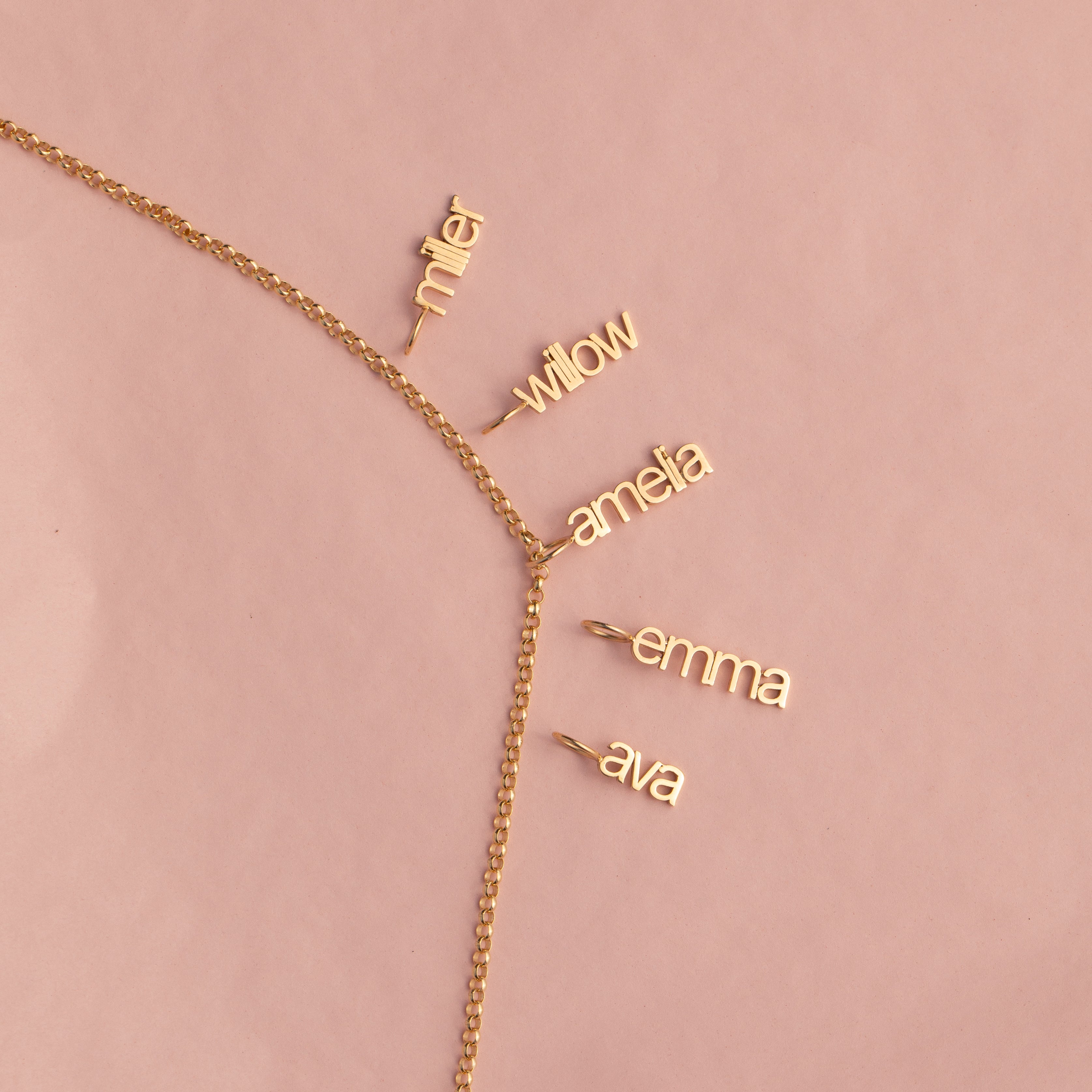 A delicate gold chain necklace laid flat on a dusty pink background, featuring five small dangling nameplates in cursive script reading 'miller', 'willow', 'amefa', 'emma', and 'ava'.
