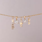 A gold chain necklace with three dangling nameplates spelling 'anabel', 'miller', and 'elva', alongside two clear round gemstones, shown against a solid light gray background.
