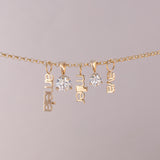 A gold chain necklace with three dangling nameplates spelling 'anabel', 'miller', and 'elva', alongside two clear round gemstones, shown against a solid light gray background.